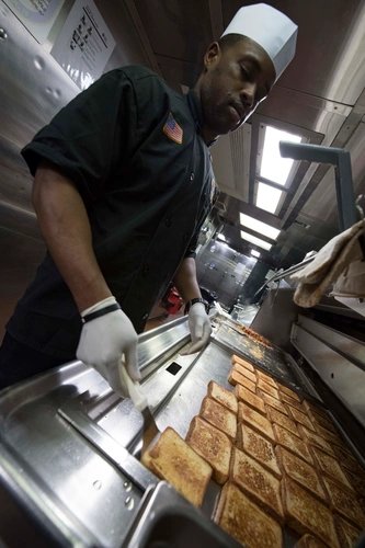 Chef Moe, in a Navy chef's hat and uniform, preparing grilled sandwiches on a griddle in a U.S. Navy ship's galley.