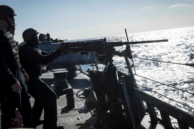 Chef Moe and fellow sailors on watch at a .50-caliber machine gun mount on the deck of a U.S. Navy ship, backlit by sunlight on the open ocean.